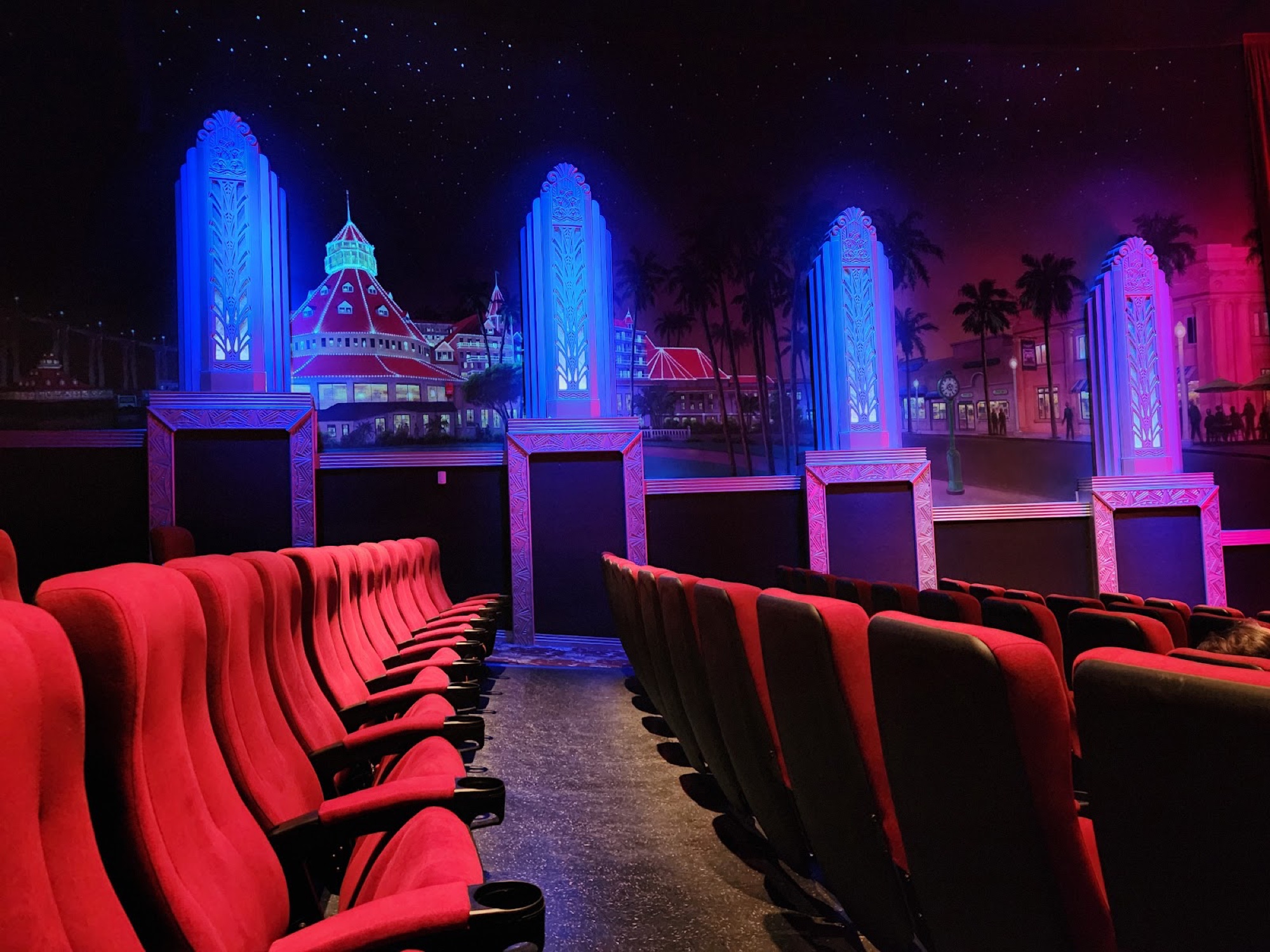 Atmospheric Village Theatres auditorium in Coronado: red velvet seats facing an Art Deco proscenium painted with the floodlit Hotel del Coronado and palm-lined village under a starry indoor sky